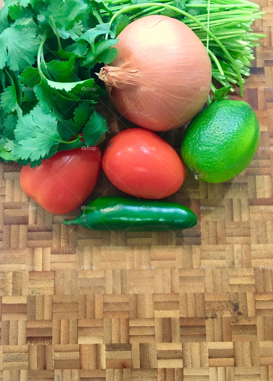 Cilantro, onion, tomato, jalapeño, and a lime placed on top of of wooden cutting board.