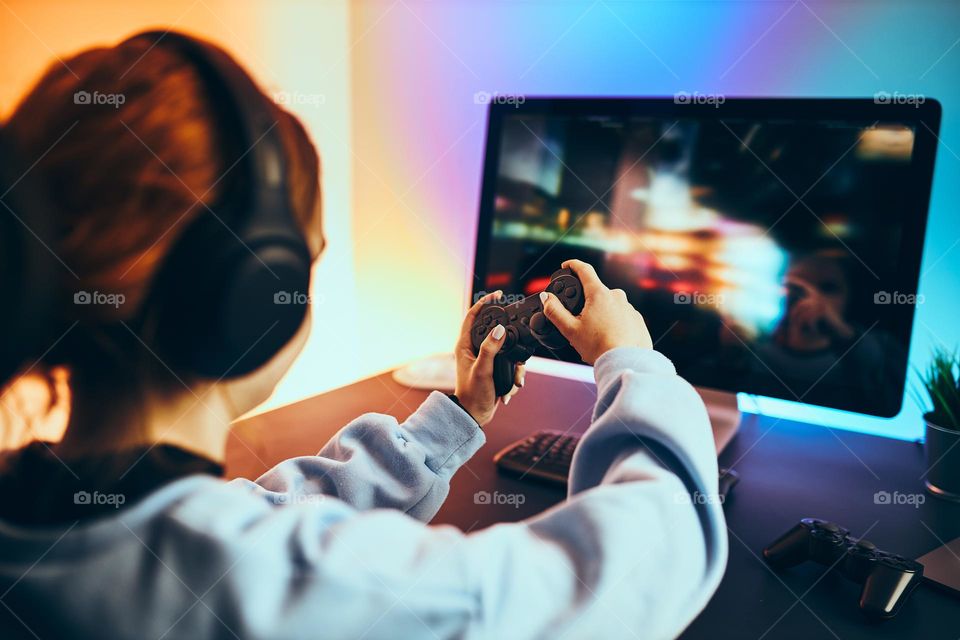 Teenager girl playing video game at home. Gamer holding gamepads sitting at front of screen. Streamer playing online in dark room lit by neon lights. Competition and having fun