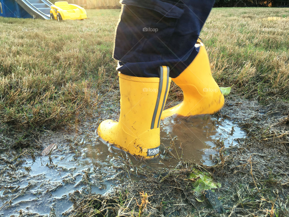 Yellow totes rain boots in backyard mud puddle. Child playing in water wearing yellow rain boots