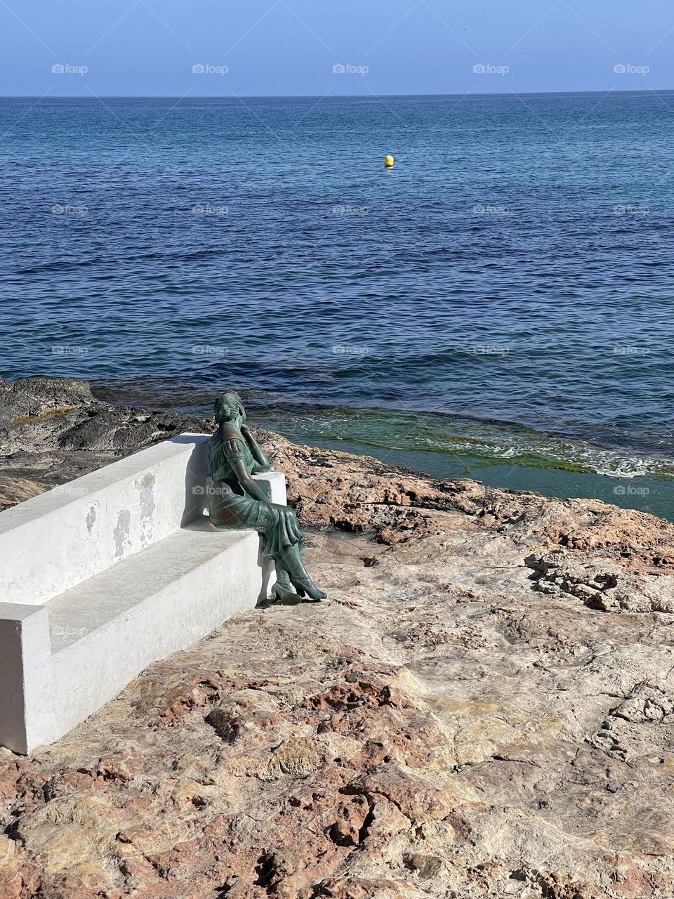 Statue of woman looking at the sea sitting on a white stone bench 