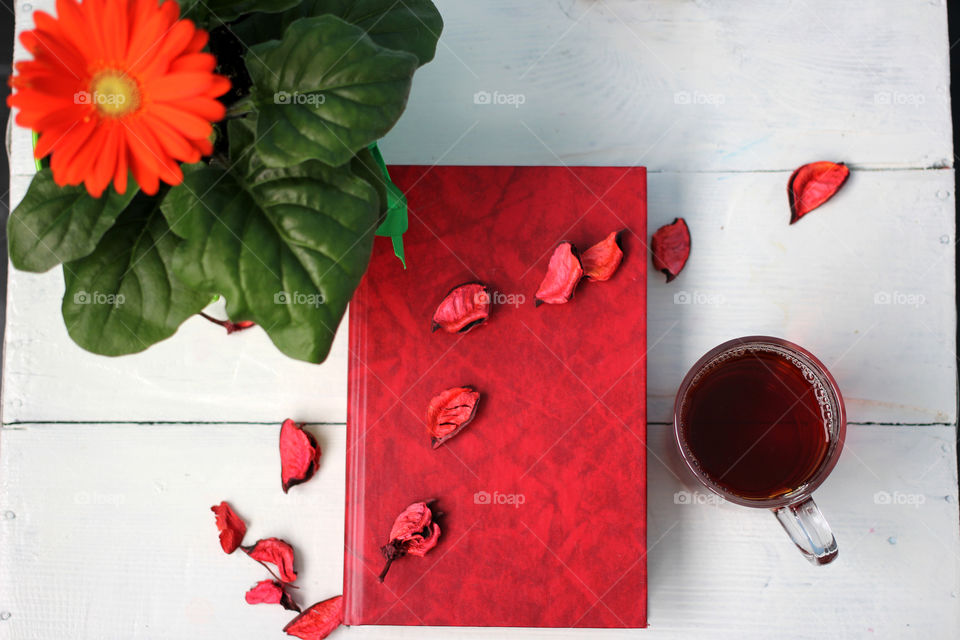 Flower with a cup of tea and a book on a white table, which is decorated with rose petals