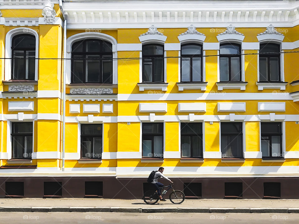 A cyclist using a mobile phone and riding a bicycle in front of the old yellow geometric building 