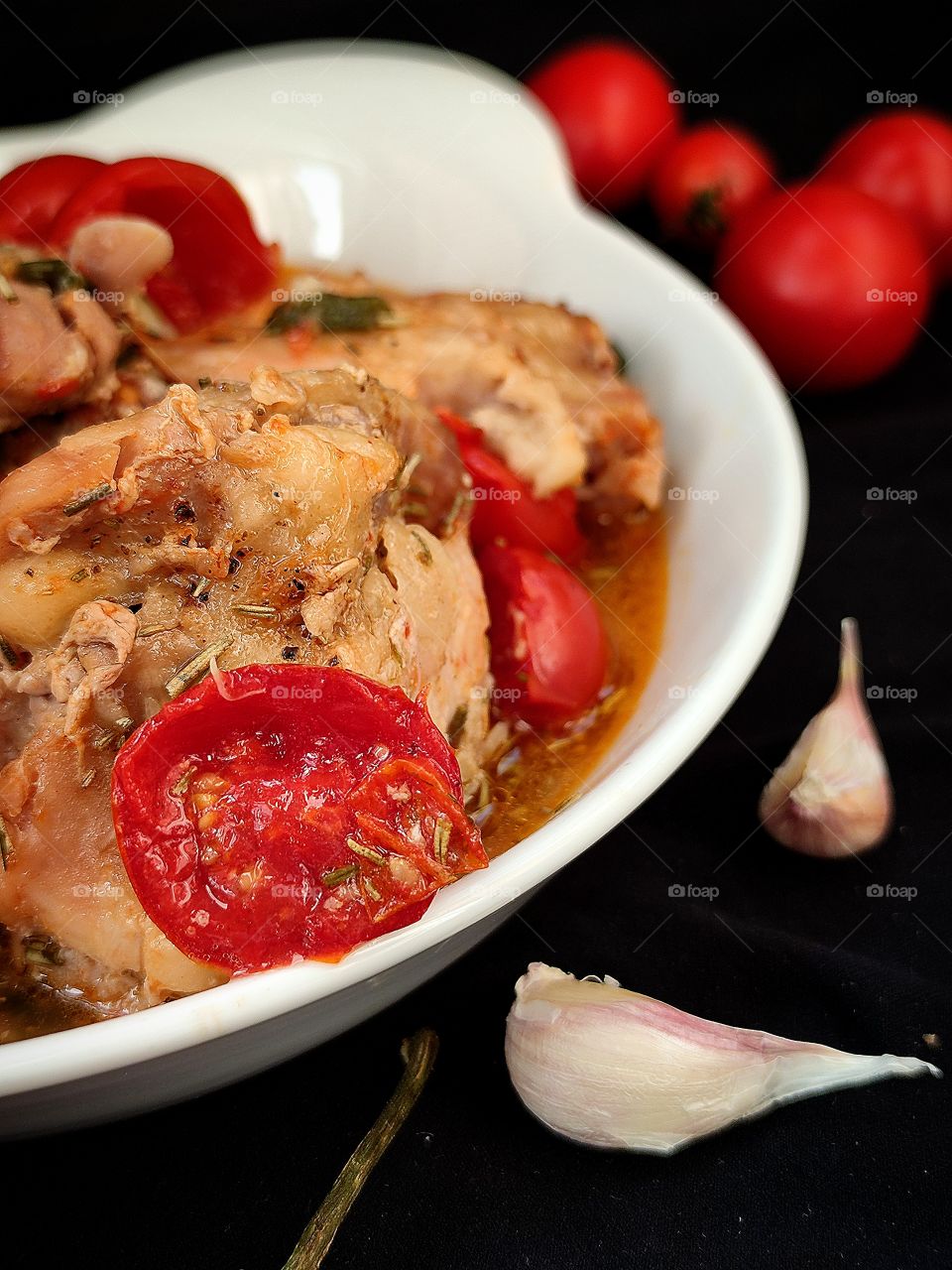 stewed meat with spices and cherry tomatoes in a white plate.  Nearby are two cloves of garlic and cherry tomatoes.  Black background