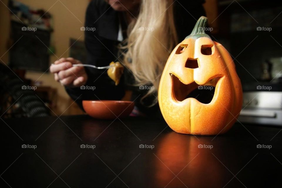 A Jack O'Lantern stands on a black table in the kitchen and a woman is eating in the background