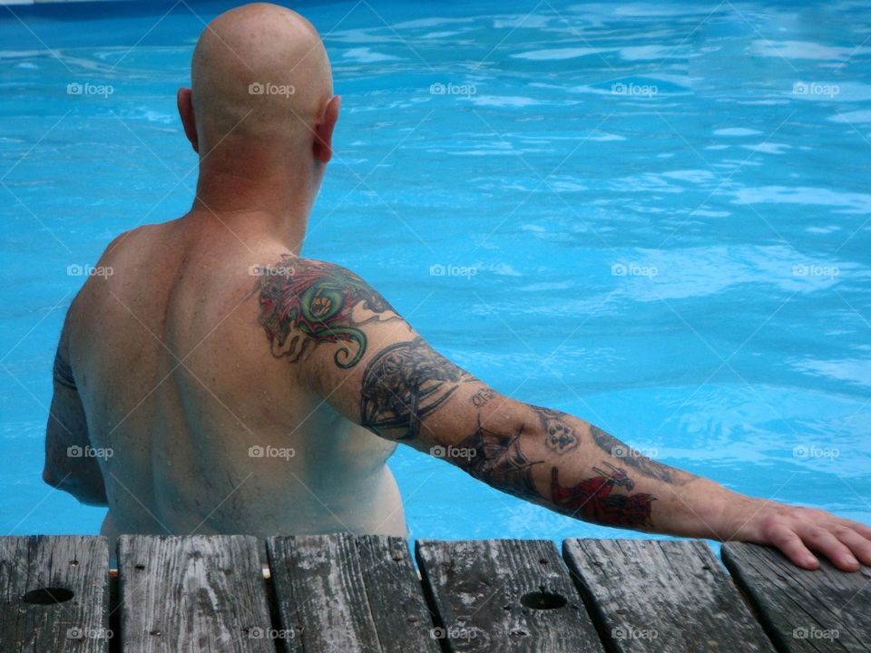 Man standing waist deep in backyard swimming pool. His back is turned & he has an arm tattoo as he's sunbathing.