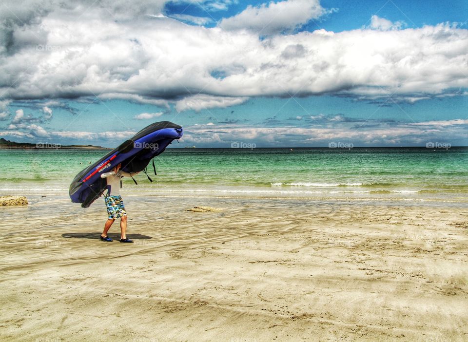 Carrying A Boat. A boy carrying an inflatable dinghy on his head along a sandy beach.