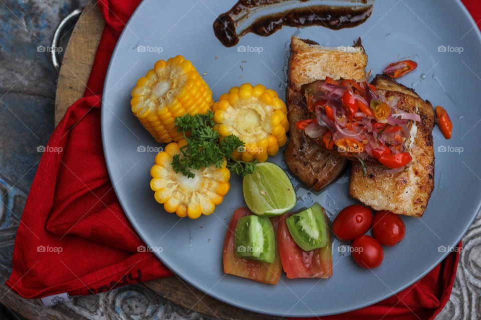 Tuna steak cooked with corn garnished with cherries, sliced tomatoes and lemon topped with sliced hot chili peppers on a baby blue melamine plate lined with red cloth and thick wood. Restaurant food.