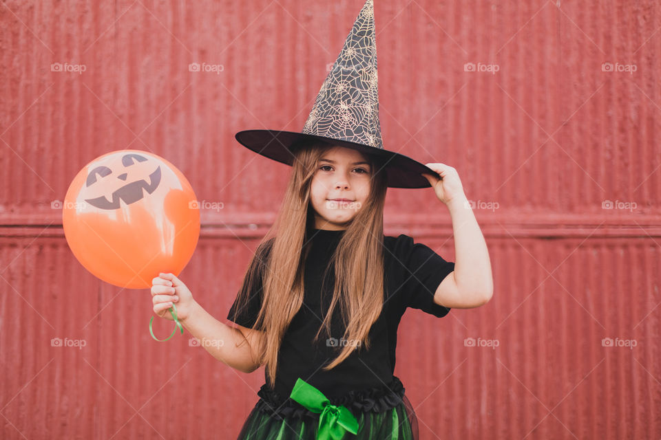A little girl dressed as a witch for halloween holds a pumpkin-shaped balloon in her hands.