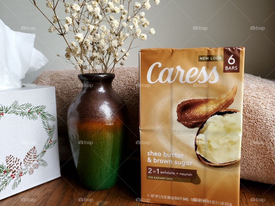 The new look Caress shea butter and brown sugar six pack bar soap on a wood surface with a flower vase, tissue box and tan towel.
