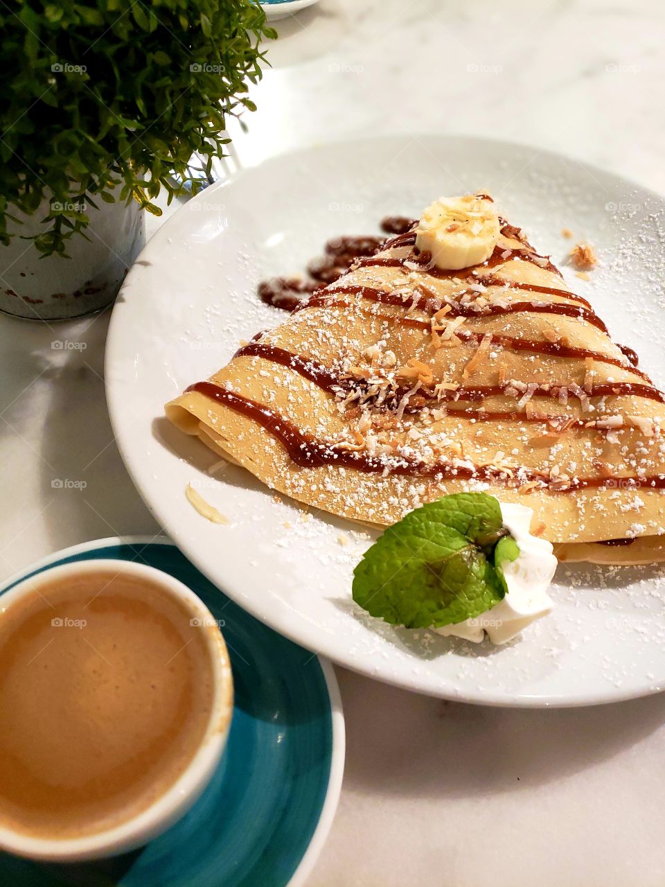 Crêpe with banana and drizzled caramel sauce next to a latte and potted herb plant.