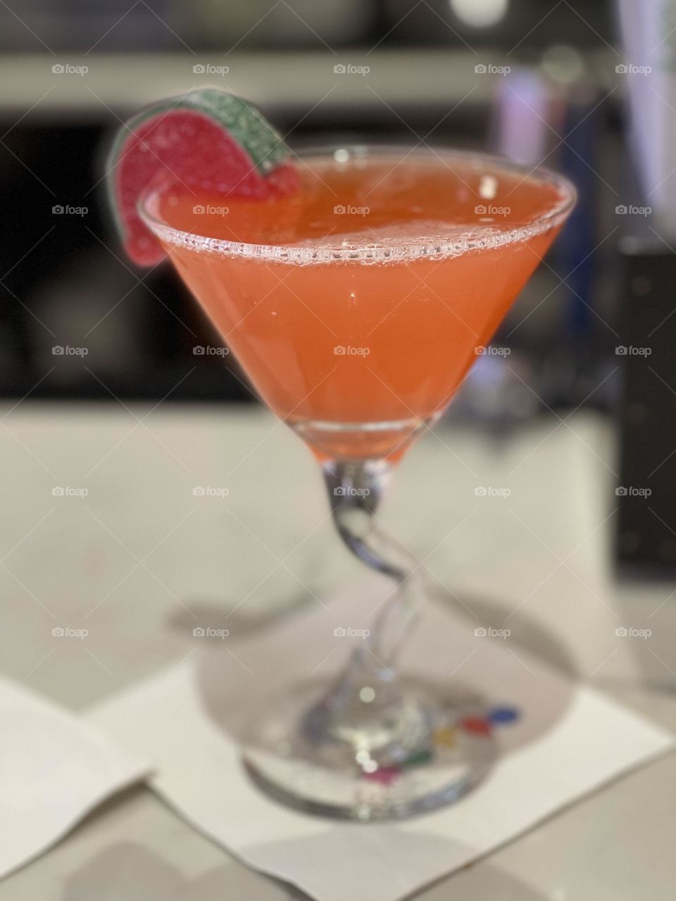 A strawberry martini is served in a funky glass with a gummy on the rim. The drink sits on a candy bar ready to be enjoyed.