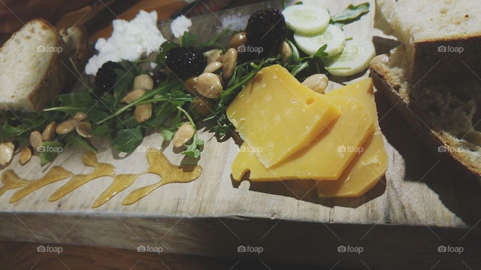 Assortment of cheese and fruit with honey, bread, and herbs on a multi colored wooden tray.