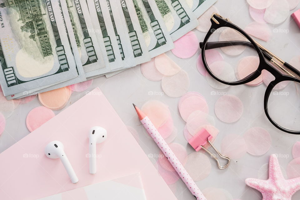 Flatlay items: cash money one hundred dollars bill, reading glasses, wireless headphones, notebook, pencil, star pink color, confetti lying on gray concrete background 