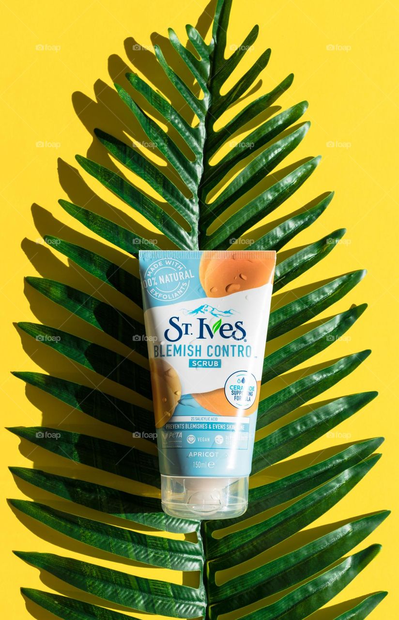 One tube of peach scrub brand St. Ives lies on a green palm leaf in the middle on a yellow background, close up.