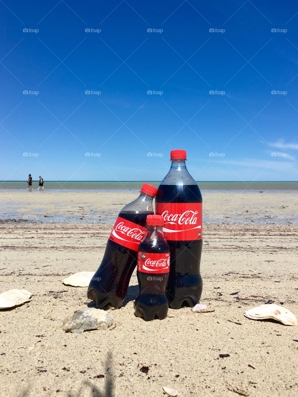 A “Family” (think mama, papa, and the little one) represented by three bottles of varying sizes of Coca Cola, spending time at the beach. See the family in the background on the ocean horizon 