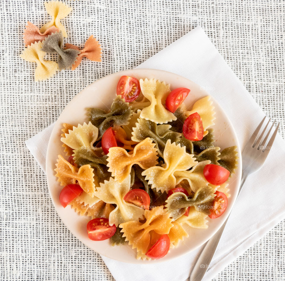 Top view of italian colored pasta farfalle with basil and tomatoes on the light plate at the table.
