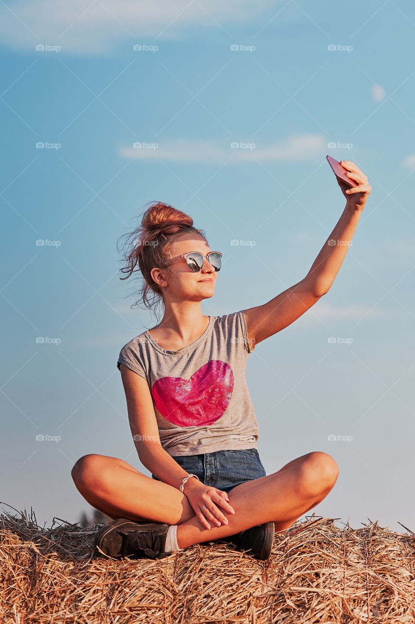 Teenage girl taking a selfie of herself, sitting on a hay bale enjoying summer vacation in the countryside. Candid people, real moments, authentic situations