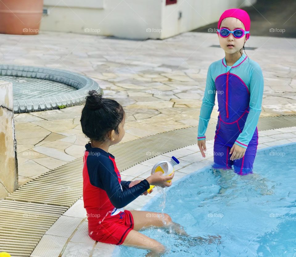 Two beautiful little girls are wear swimming outfits, one of them has pink cap on her head and both playing at swimming pool 