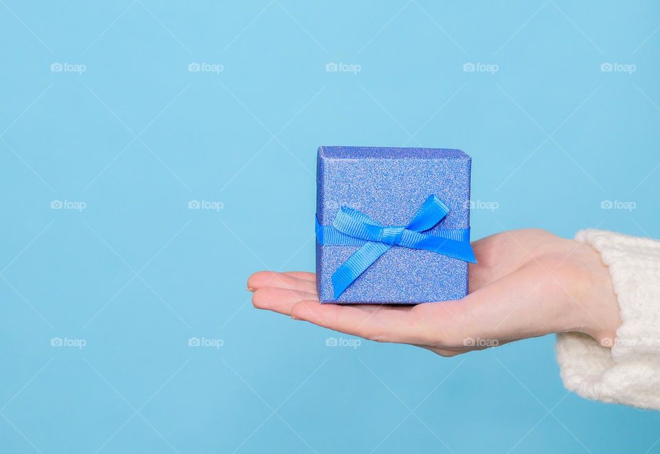 The hand of a young caucasian girl is holding in the palm of her hand a small blue shiny gift box on the right on a wall background with copy space on the left, close-up side view. Holiday gifts concept.