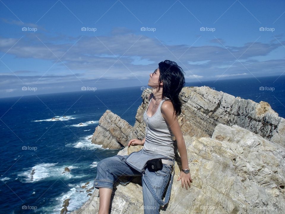 It's a girl sitting on a giant rock on the beach. Behind you see the blue sea and the sky. The photograph is made in horizontal.