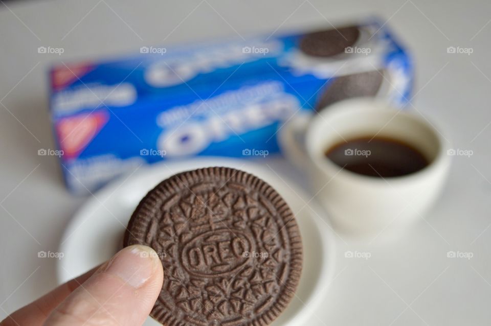A persons fingers holding an Oreo cookie and a cup of coffee in the background