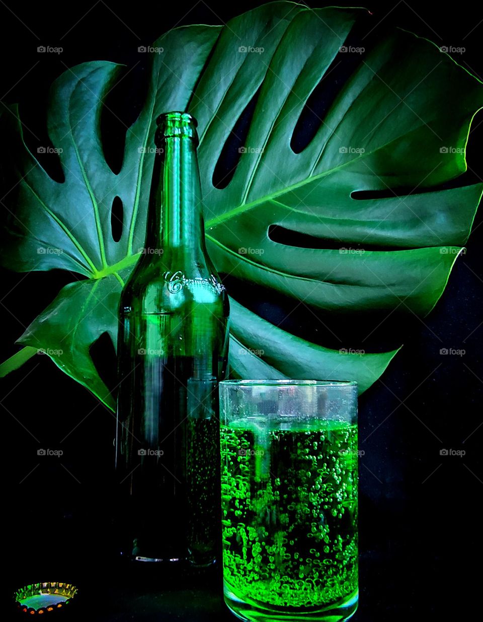 Green color.  On a black background, a green leaf of Monstera, a green glass bottle and a glass with a green drink. There is a metal cork next to the bottle.