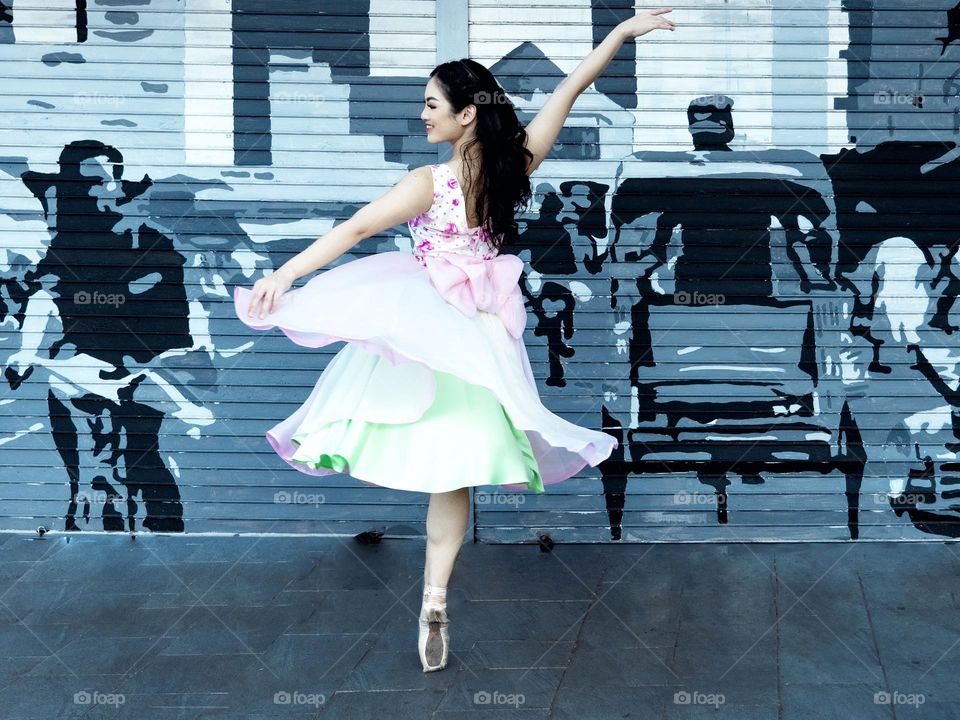 A ballerina is dancing by the side of the road, with a background to a mural that shows the life around.