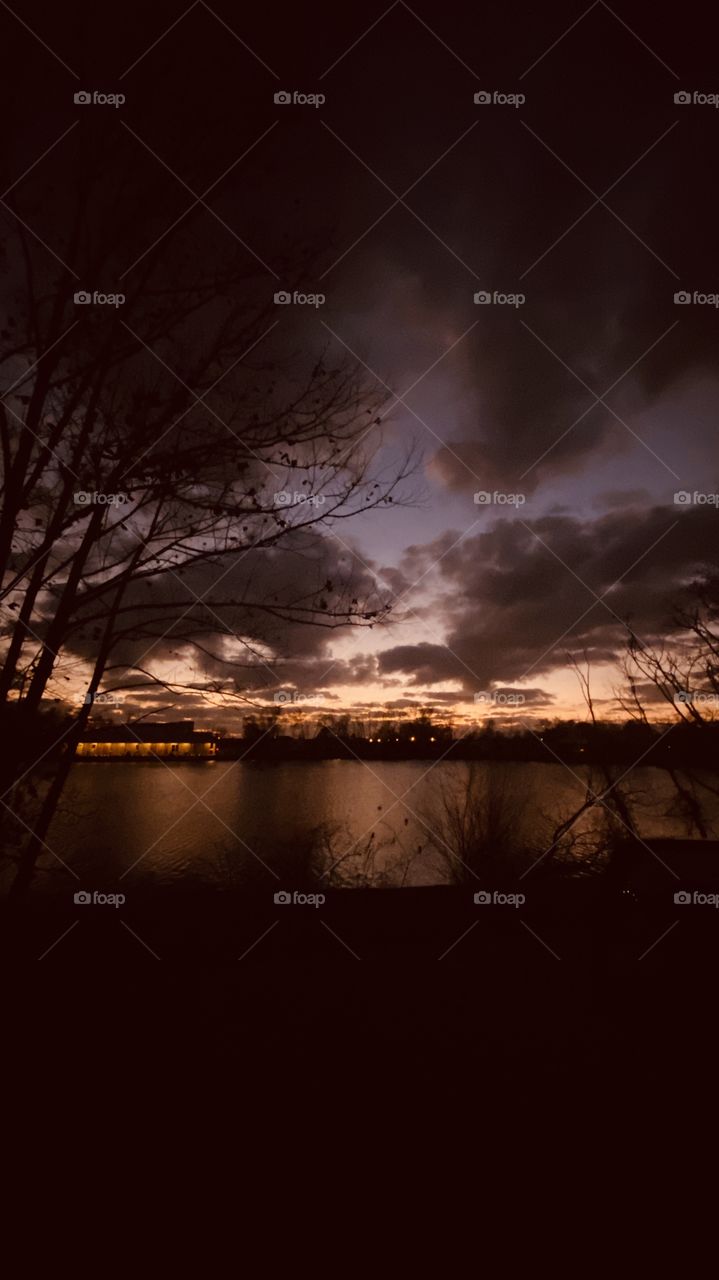 Sunset-Twilight-Night Visible Backlit Skyline. Lake Water Reflections of ShoreLine to Calm Lake Waters. Reflections of Hanging Clouds off Lake