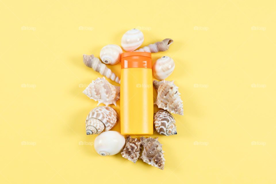 One small yellow bottle with an orange cap of suntan cream lies in the center on a light yellow background, and sea shells are located around, flat lay close-up.