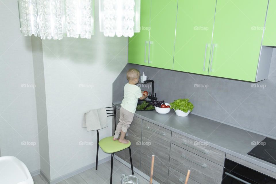 A small child washes fresh, green, vegetarian vegetables under the tap in a black sink to prepare salads and other dishes in a homemade gray and green kitchen.