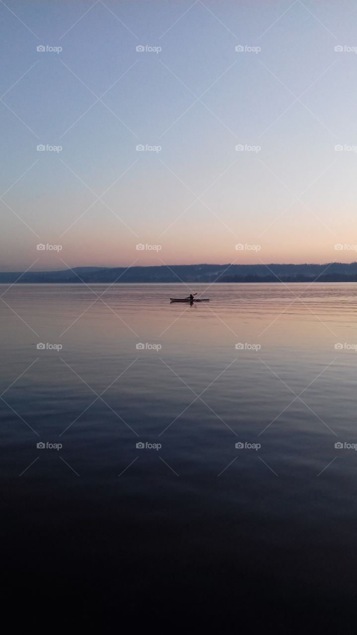 A large lake photographed at sunset with a far small canoe in the center, all the sky is coloured by blue and orange