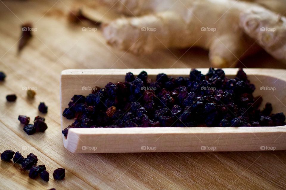 Closeup of a wooden scoop full of dried elderberries on a wooden surface with scattered elderberries and whole cloves, ginger root in the background