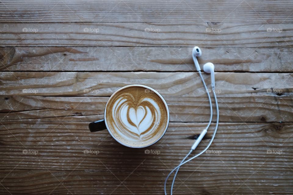 A cappuccino with latte art sitting next to a pair of Apple ear bud headphones on a table in the window light. 