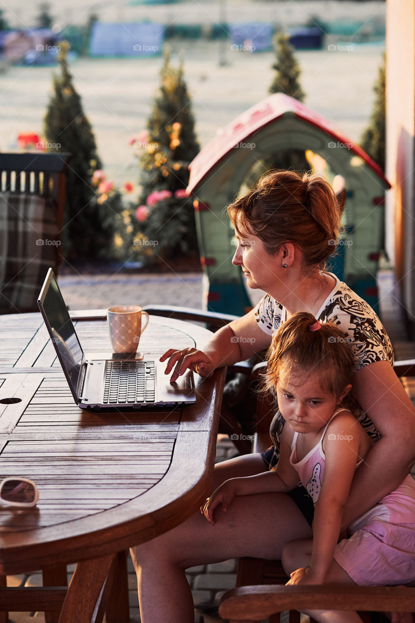 Mother working at home, using portable computer, sitting on patio with her daughter on summer day. Candid people, real moments, authentic situations