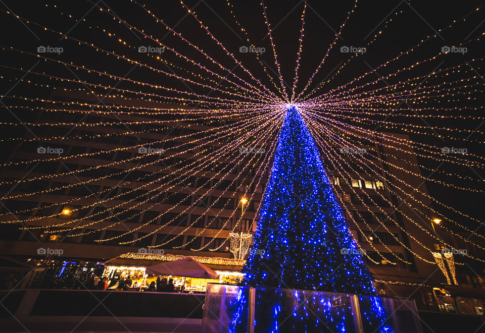 Outdoor Illuminated Blue Christmas Tree With Blue Lights At Square With  People Enjoying Holidays
