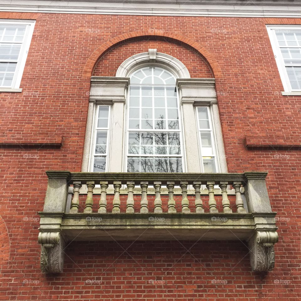 This image looks up at an ornate window painted white set in a large red brick wall. There is a decorative cement balcony Beneze the window. The window is tall and has a semi circular top.