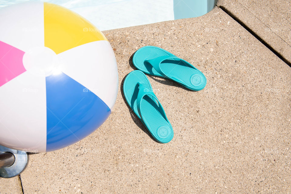 Overhead view of a beach ball and women's flip flops next to a swimming pool