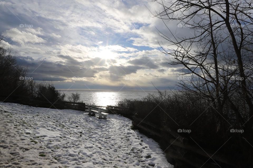 Winter by the ocean , snow covered ground and empty bench against dramatic sky