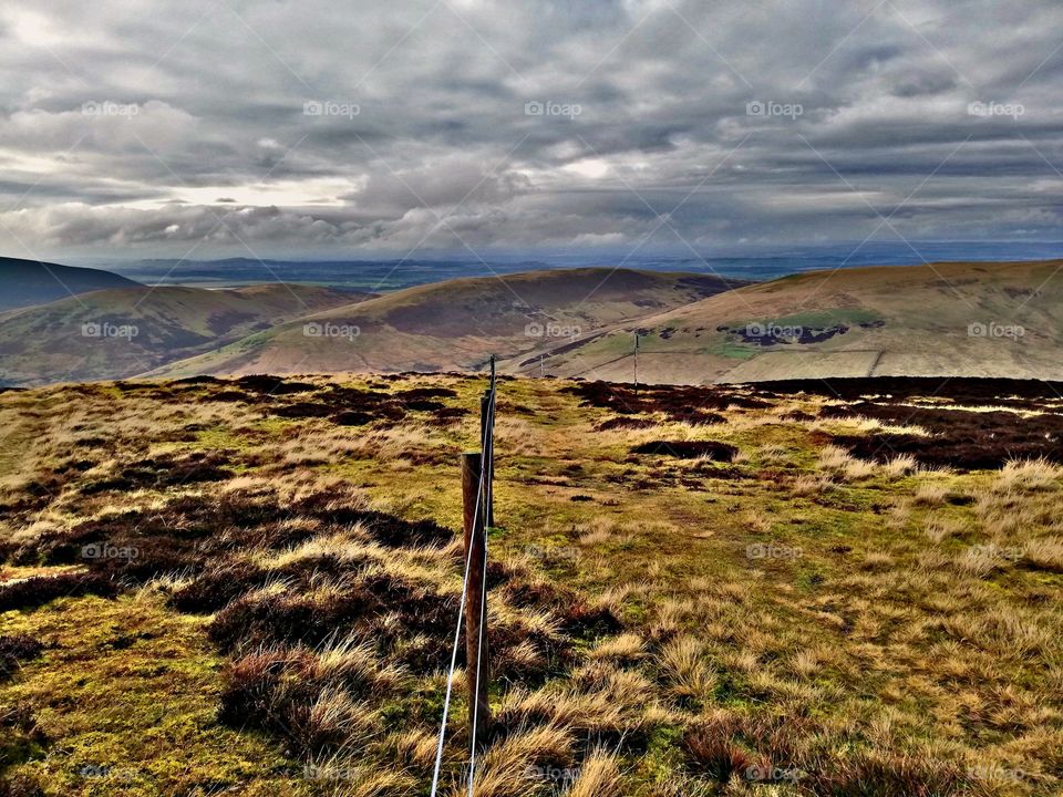 a fence line on top of a hill in Scotland. u can see the cloud's in the sky with a bit a bluesky in the background. look at the fence in the distance with the beautiful grass