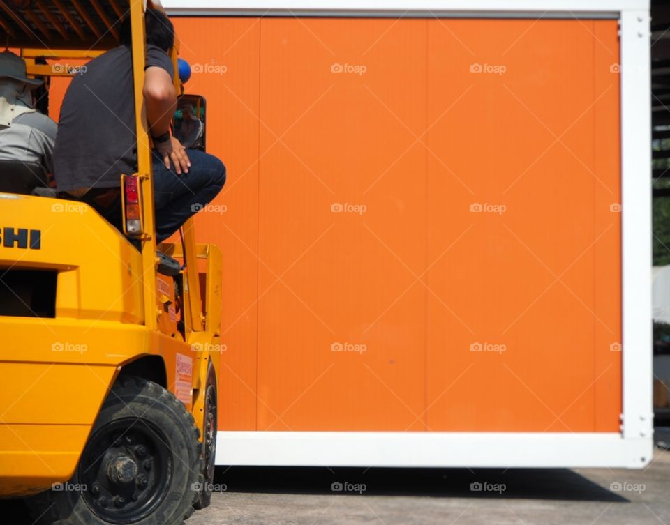 An driver and his assistant is sitting on top of the forklift while they are lifting a container.