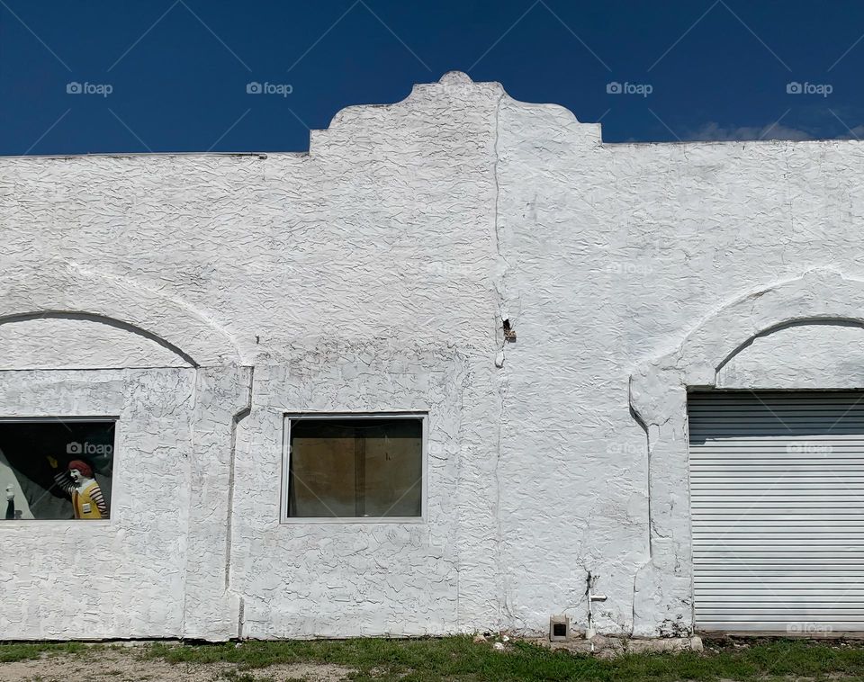 Old white architectural building with signs of aging elements under a blue sky in the afternoon with turned edges arched door and window with a peaking character on the left window.