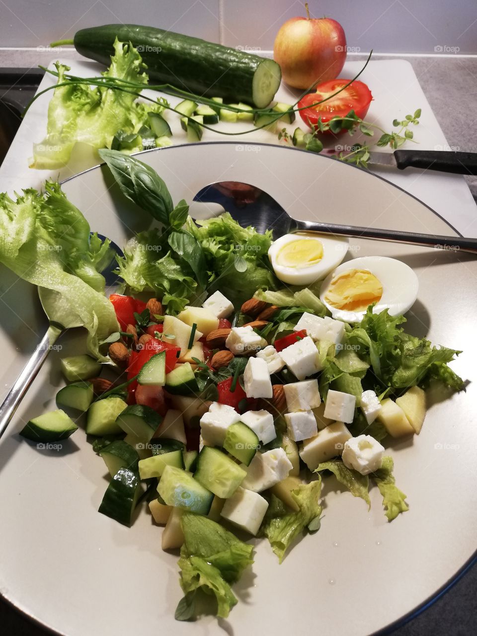 Delicious salad in the bowl with the handles. Lettuce, cucumber, tomato, cheese, almond, boiled egg, apple, chives, thyme, basil and salad dressing. In the background some ingredients on the chopping board.