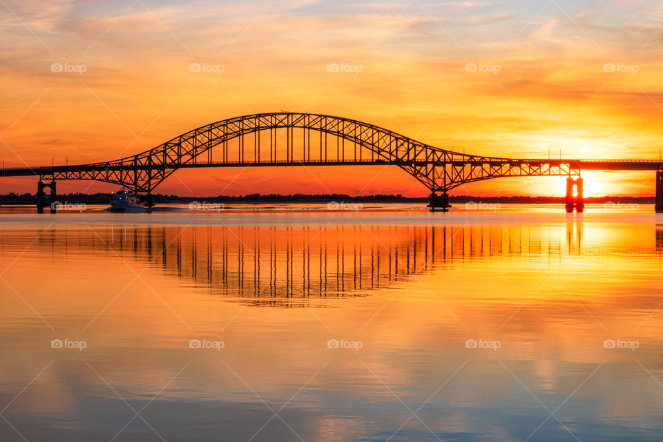 Silhouette of a steel tied arch bridge crossing a large bay, with a colorful vibrant sunset reflecting in the water. 