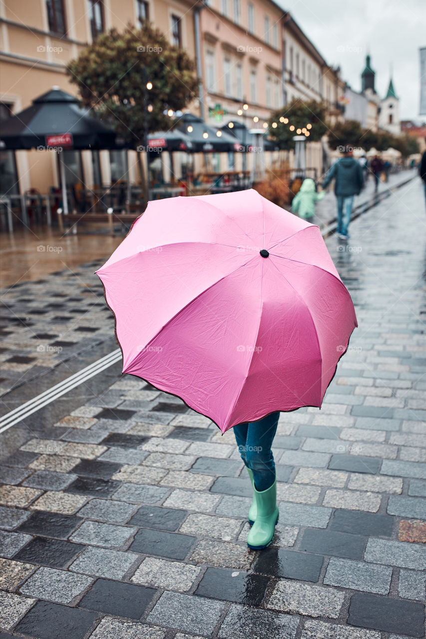 Little girl hiding behind big pink umbrella walking in a downtown on rainy gloomy autumn day