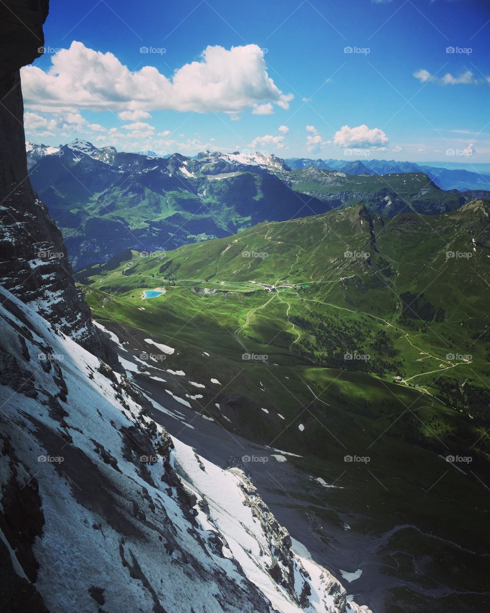 I am standing atop of summer snowy mountains, black mountain and white snow. It is surrounded by green mountains, and pathways can be seen there, there is a very beautiful blue lake, The blue sky has large white clouds, beautiful sunny day