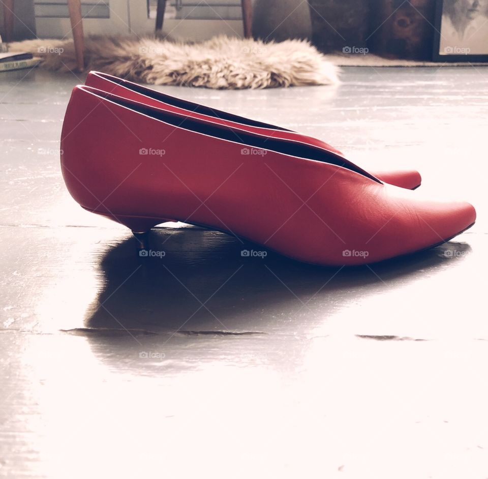 Red leather kitten heeled pump shoes on painted grey floorboards with sheepskin rug in the background. Picture taken in natural daylight