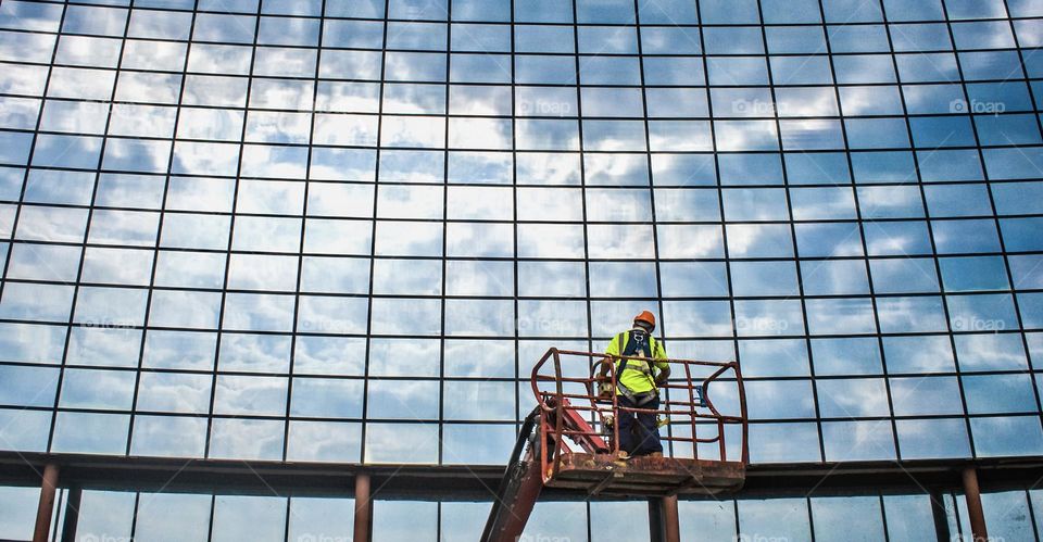 Man in high vis & hard hat is held in a lifting cradle in front of dozens of window reflecting a blue sky with clouds