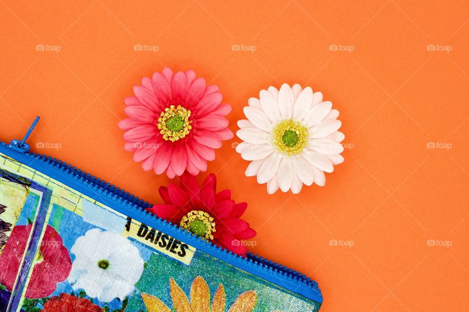 Flat lay of Gerbera daisies and a colorful floral-print zipper bag with blue trim on a bright orange background 
