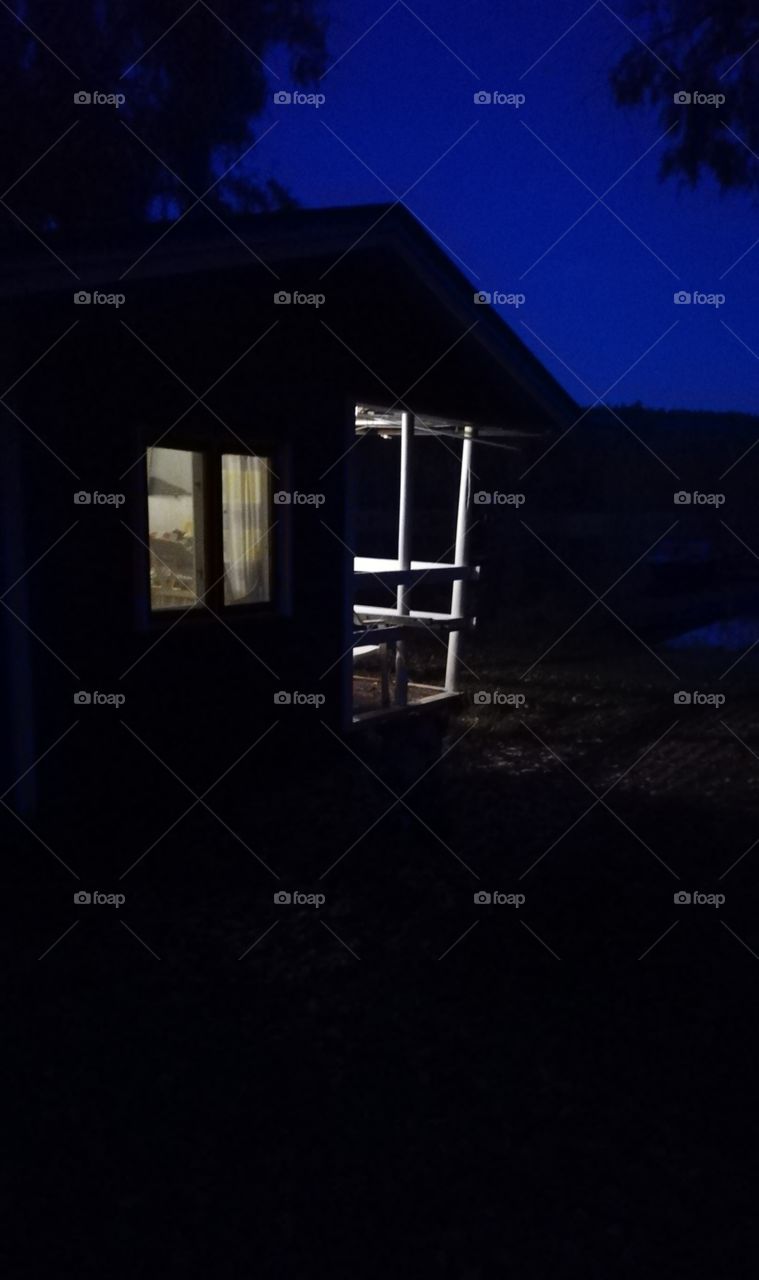 A lake side sauna building in the dark autumn evening. The bright light gives shadows on the ground and the window and wooden parts of the terrace are clearly seen. The reflection of the blue sky on the lake.