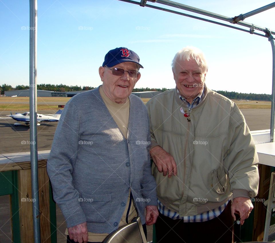 Two older men at outside restaurant overlooking small plane airport. Best friends, senior citizens, happy, smiling, one uses a cane. Small airplane in background.